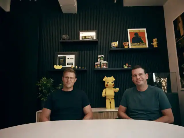 Two men sit at a round table in an office with black walls, shelves displaying awards, books, and Pikachu-themed items behind them.